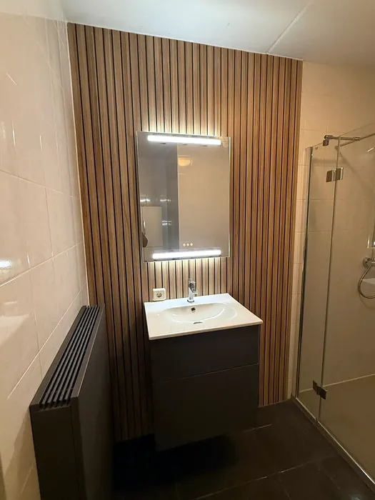 Badkamer met glazen douchecabine, hangtoilet tegen een lamellenwand en donker meubel